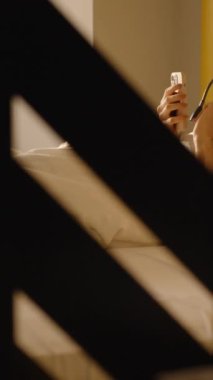 Dikey video. Beyaz bir yorganın altında, sarı aksanlı ve modern yatak odasıyla çevrili, sabah güneşinde akıllı telefonunu kullanan bir kadın..