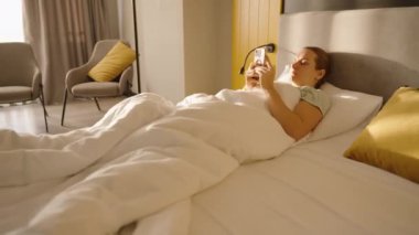 Bir kadın, beyaz bir yorganla yatakta uzanıyor. Telefonunu sarı ve gri aksanlı, parlak ve modern bir yatak odasında kaydırıyor. Huzurlu bir sabah havası yakalıyor..