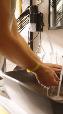 Dikey video. Modern bir banyo lavabosunda ellerini parlak bir musluğun altında yıkayan bir kadın. Yumuşak ışıklandırma ve su damlacıkları hareket halindeyken, hijyen ve temizliği ön plana çıkarıyor..