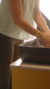 Dikey video. Siyah, modern bir lavaboda ellerini yıkayan bir kadının yan görüntüsü. Parlak bir banyoda, sakin ve temiz bir anı yakalıyor..