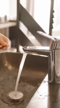 Dikey video. Çağdaş bir banyoda temizlik ve temizliği vurgulayan su damlacıkları ve ışık yansımalarıyla ellerini parlak bir musluğun altında yıkayan bir kadının yakın çekimi..