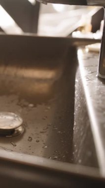 Dikey video. Su damlacıklı parlak siyah bir lavabo ve modern bir musluk çağdaş bir banyoda duruyor. Arka plandan ışık akıyor ve sakin bir atmosfer yaratıyor..