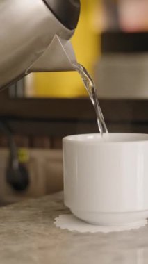 Dikey video. Su ısıtıcısı, mermer tezgahın üzerindeki beyaz bir fincana sıcak su döker. Arka planda cam eşyalar ve yansımalar, sakin ve rutin bir anı yakalar..