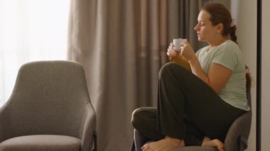 Bir kadın pencerenin yanındaki sandalyeye kıvrılmış, beyaz bir bardaktan sıcak bir içkinin tadını çıkarıyor, yumuşak doğal ışıkta rahat ve huzurlu bir atmosfer yaratıyor..