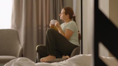 Bir kadın pencerenin yanındaki sandalyeye kıvrılmış, elinde beyaz bir bardakla oturur, rahat bir ortamda perdelerden süzülen doğal ışıkla sakin bir sabahın tadını çıkarır..