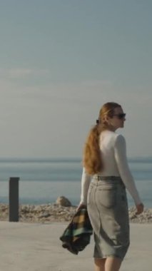 Dikey video. Bir kadın, elinde ekose bir ceketle, sakin bir deniz kıyısında yürürken berrak bir gökyüzü altında, sakin bir kıyı sahnesi yaratıyor..