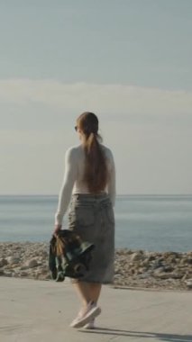 Dikey video. Bir kadın berrak mavi bir gökyüzünün altında sakin bir sahil boyunca yürüyor, ekose bir ceket giyiyor ve huzurlu kıyı manzarasının tadını çıkarıyor..
