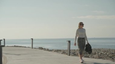 Bir kadın, açık mavi gökyüzü ve parlak güneş ışığı altında, elinde ekose bir ceket ile, deniz kenarındaki gezinti alanında güvenle dolaşır..