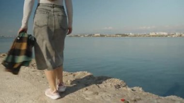 Kayalık bir sahil yolunda yürüyen, elinde ekose ceketi olan, arka planda açık mavi deniz ve gökyüzü olan bir kadının yakın görüntüsü..