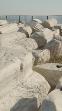 Dikey video. Parlak mavi gökyüzünün altında denize bakan korkulukların yanındaki tarihi kalıntılar, dağınık antik mermer bloklardan oluşan bir koleksiyon..