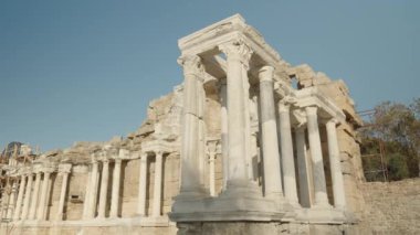 Yüksek beyaz mermer sütunları, detaylı oymaları ve yıpranmış taş duvarları olan, parlak mavi bir gökyüzünün altında alçak bir açıyla çekilmiş antik bir Roma yapısı..