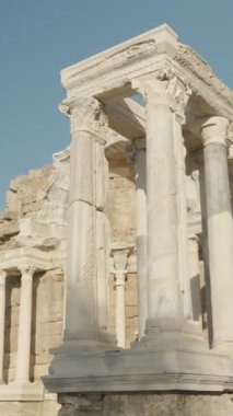 Dikey video. Yüksek beyaz mermer sütunları, detaylı oymaları ve yıpranmış taş duvarları olan, parlak mavi bir gökyüzünün altında alçak bir açıyla çekilmiş antik bir Roma yapısı..