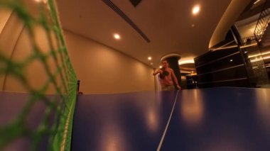 Yakın çekim, düşük açılı bir masa tenisi maçı görüntüsü, masa ve ağa odaklı, kapalı bir odada sıcak ışıklandırmalı..