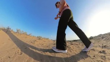 Açık mavi gökyüzünün altında kuru bitki örtüsüyle kumlu arazide yürüyen ve gölgeyi ve doğal ortamı yakalayan bir kadının düşük açılı perspektifi..