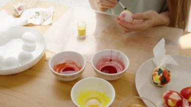 Kadın mermer pembe ve sarı bir Paskalya yumurtası boyamak için fırça kullanıyor. Masanın yanında küçük bir kavanoz sarı boya var..