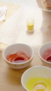 Dikey video. Kadın mermer pembe ve sarı bir Paskalya yumurtası boyamak için fırça kullanıyor. Masanın yanında küçük bir kavanoz sarı boya var..