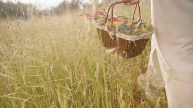 Bir kadın uzun otların arasında yürürken bir sepet renkli Paskalya yumurtası ve çiçek taşır..