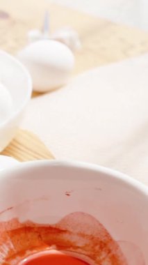 Dikey video. Paskalya yumurtasını paspasın üzerine yuvarlayan bir kadın. Yanında bir kase boya ve beyaz yumurta var. Canlı Paskalya süslemeleri yapıyor..