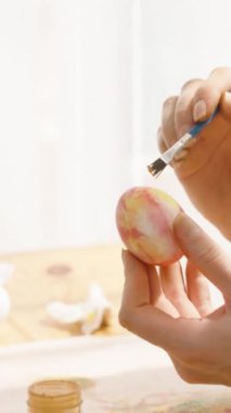 Dikey video. Paskalya yumurtasının parlak, güneşli bir odada altın aksanlı resmini dikkatlice yapan bir kadın. Karmaşık detaylar ekliyor..