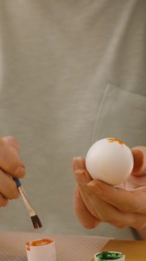 Dikey video. Paskalya yumurtasını parlak, güneşli bir odada küçük bir boya fırçasıyla süsleyen kadın, tabakta tamamlanmış renkli yumurtalarla çevrili..