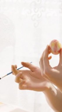 Dikey video. Aydınlık, güneşli bir odada Paskalya yumurtası üzerine hassas desenler çizen odaklanmış bir kadın. Masada çeşitli dekorasyon malzemeleri var..