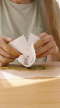 Dikey video. Paskalya yumurtasını peçeteye saran kadın, güneş ışığı parlak odaya akıyor..