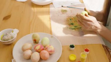 Sarı bir Paskalya yumurtası tutan kadın, parlak bir odada boyalı yumurta ve boya kavanozlarıyla çevrili..