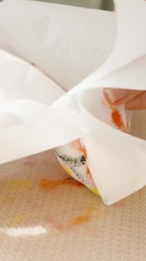 Dikey video. Renkli bir Paskalya yumurtasını bir bardak suya sarıp masaya bir kase boya ve boya malzemesi koyuyor..