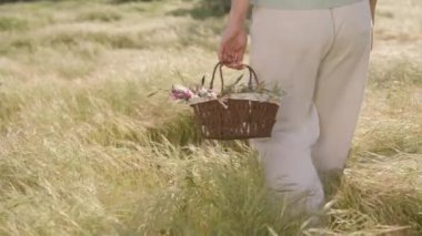 Beyaz pantolonlu bir kadın, elinde renkli çiçekler ve paskalya yumurtalarıyla dolu bir sepet tutarak güneşli bir arazide yürüyor. Yavaş çekim.