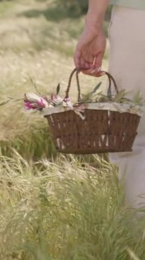 Dikey video. Beyaz pantolonlu bir kadın, elinde renkli çiçekler ve paskalya yumurtalarıyla dolu bir sepet tutarak güneşli bir arazide yürüyor. Yavaş çekim.