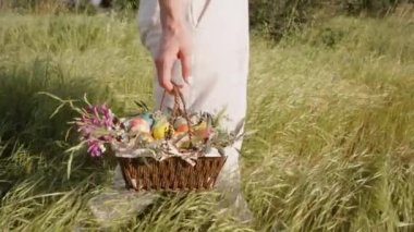Çiçeklerle Dolu Dokuma Sepeti ve Paskalya Yumurtaları Beyaz Pantolon Giyen Uzun Çimenler İçinde Yürüyen Kadın tarafından taşınır. Yavaş çekim.