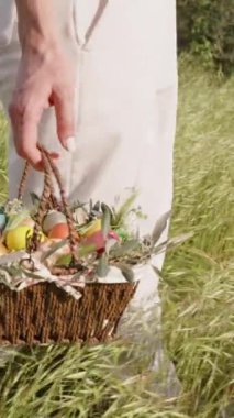 Dikey video. Çiçeklerle Dolu Dokuma Sepeti ve Paskalya Yumurtaları Beyaz Pantolon Giyen Uzun Çimenler İçinde Yürüyen Kadın tarafından taşınır. Yavaş çekim.