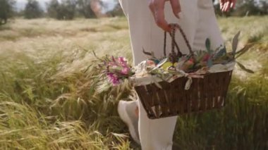 Beyaz pantolonlu bir kadın elinde çiçeklerle dolu bir sepet ve paskalya yumurtası tutarak güneşli bir arazide yürüyor. Yavaş çekim.