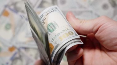 Arka planda dağınık dolar banknotları olan yüz dolarlık banknotları çeviren bir elin yakın çekimi hareket ve zenginlik hissi yaratıyor.