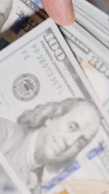 Dikey video. Yüz dolarlık banknotların el ele tutuşması ve fırlatılması, bulanık arka planda dağınık bir para yığını, hareket ve bolluğu vurguluyor.