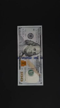 Dikey video. Karanlık bir arka plan karşısında havada süzülen tek bir yüz dolarlık banknot. Çerçevenin dışına sürüklenirken, geçici zenginliği veya fırsatı sembolize ederek hareket ederken yakalandı.