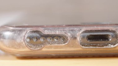 Akıllı telefonun şarj portunun ve toz ve molozla kaplı hoparlör deliklerinin yakın çekimleri, kirliliği ve ihmali vurgulamak için sürgülü hareketle yakalanır.