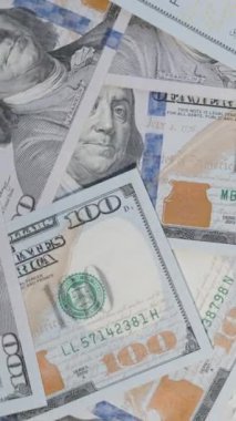 Dikey video. Dağınık yüz dolarlık banknotların hava görüntüsü, döndüklerinde düzenlemelerini ve hareketlerini gösteriyor, finansal akışı ve parasal bolluğu vurguluyorlar.