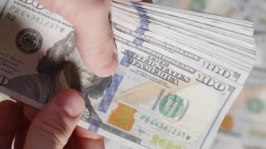Kalın bir yığın 100 dolarlık banknotu tutan bir elin yakın plan görüntüsü, doğal ışık altında paranın dokusunu ve detaylarını vurguluyor.