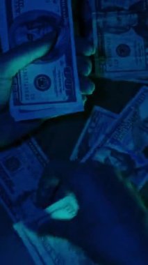 Dikey video. Bir kadın aceleyle mavi ışığın altında dağınık bir masanın üzerindeki yığından Amerikan doları banknotları topluyor, aciliyet ve gerilimi iletiyor.
