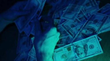 Bir kadın, hüzünlü mavi ışıklandırma altında, parayı sınıflandırmaya ve idare etmeye odaklanmış, dağınık Amerikan doları banknotlarını yakından inceleyip düzenliyor.