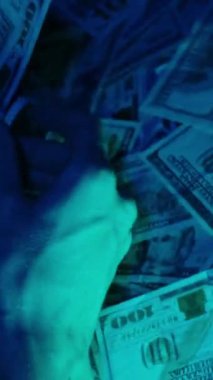 Dikey video. Bir kadın, hüzünlü mavi ışıklandırma altında, parayı sınıflandırmaya ve idare etmeye odaklanmış, dağınık Amerikan doları banknotlarını yakından inceleyip düzenliyor.