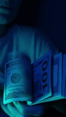 Dikey video. Yakın plan çekimde dramatik mavi ışıklandırma altında bir yığın Amerikan doları banknotunu sayarken masanın üzerinde dağınık banknotlar görülebiliyor, gergin ve odaklanmış bir an yaratıyor.