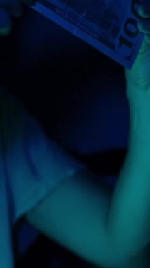 Dikey video. Bir kadın, yumuşak mavi ışık altında yüz dolarlık banknotu inceliyor. Odaklanmış ifadesi dikkatle incelendiğini veya doğrulandığını gösteriyor.