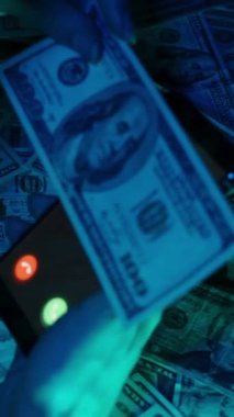 Dikey video. Gelen dolandırıcılık ihbarını gösteren bir akıllı telefon, dramatik mavi ışıklandırmayla aydınlatılan ve finansal dolandırıcılığı vurgulayan Amerikan doları banknotlarıyla dolu bir masanın üzerinde tutuluyor.