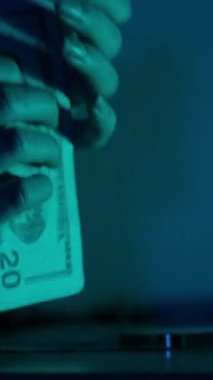 Dikey video. Huysuz mavi ışıklandırmanın altındaki bir masaya Amerikan doları banknotlarını istifleyerek ve düzenleyerek finansal organizasyon ve kontrol üzerine odaklanan bir kadının ellerine yakın çekim