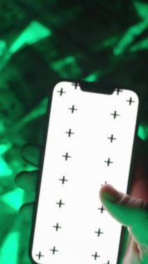 Dikey video. Boş Beyaz Ekran Modelleri ve İzleme İşaretleri ile Smartphone 'u Aşağı Çeken Bir Elin Yakın Çekimi, ABD Yüz Dolarlık Faturaların Yeşil Parlak Arkaplanı Üzerinde Konumlandırılmış, Etkileşimli Teknoloji ve Hareket Temsilcisi