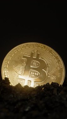 Dikey video. Karanlığın içinden çıkan Bitcoin madeni parası, Karanlık Kayalara gömülü iken ince ışığı yansıtan cilalı yüzeyi. Dijital Altın ve Madencilik kavramını simgeler