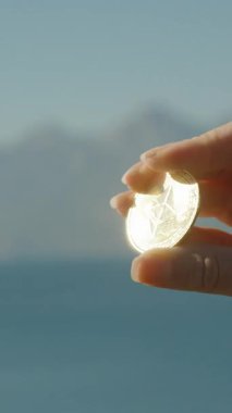 Dikey video. Mavi Okyanus 'un ve Açık Gökyüzü Olan Uzak Dağların Manzaralı Arkaplanına Karşı Parlak Bir Ethereum Parası Tutmak. Modern Dijital Finans ve Yerel Ekonominin Sembolü