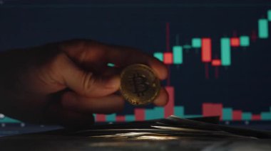 Arkaplanda Ticaret Çizelgesi olan ve Piyasa Eğilimlerini ve Mum Büyümesini Gösteren Bir Bitcoin Madeni Parası Tutmak